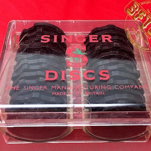 Puede incluir: Una caja de plástico transparente que contiene una pila de discos negros para máquina de coser Singer. La caja tiene el texto "SINGER DISCS" impreso en rojo en la parte delantera. El texto "THE SINGER MANUFACTURING COMPANY MADE IN GREAT BRITAIN" está impreso en negro debajo del texto rojo.