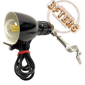 Könnte beinhalten: Eine schwarze Schreibtischlampe im Vintage-Stil mit weißem Innenschirm und klarer Glühbirne. Die Lampe hat einen flexiblen Arm und einen Klemmfuß. Das Stromkabel ist schwarz und mit einem roten Band gesichert. Die Lampe ist mit "BFTERS" beschriftet.