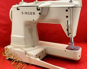 May include: Cream-colored Singer sewing machine with the brand name prominently displayed. The machine has two spool pins, a needle, and a small, pink-colored plate. The text "The Singer Company" is visible. The machine is set against a red background.