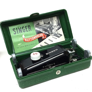 May include: A green Singer buttonholer sewing machine in its case. The case is open and the machine is visible. The machine has a black body with a silver trim and a red button. The case has a white label with the words "Singer Buttonholer" and "The Singer Manufacturing Co."