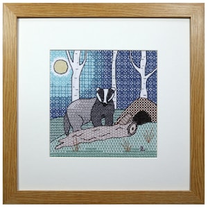 May include: A framed cross-stitch embroidery showing a badger standing near its sett in a woodland scene. The badger is grey and black with a white stripe on its face. The background is a blue and white patterned design with a full moon in the top left corner.