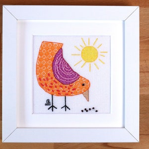May include: A framed cross-stitch embroidery depicting a stylised orange and purple bird with a yellow sun in the background. The bird is looking down at a few black stitches on the white fabric.
