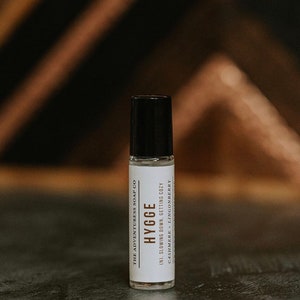 Puede incluir: Un frasco blanco de roll-on con tapa negra y etiqueta. La etiqueta dice "THE ADVENTURESS SOAP CO", "HYGGE", "(N). SLOWING DOWN, GETTING COZY", y "CASHMERE - LINGONBERRY".