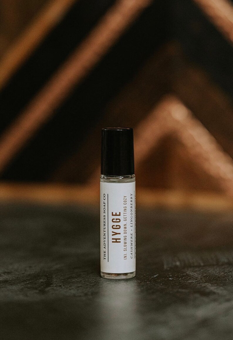 Puede incluir: Un frasco blanco de roll-on con tapa negra y etiqueta. La etiqueta dice "THE ADVENTURESS SOAP CO", "HYGGE", "(N). SLOWING DOWN, GETTING COZY", y "CASHMERE - LINGONBERRY".