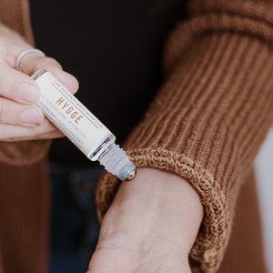 Puede incluir: Un frasco de vidrio transparente con una bola de rodillo de metal. El frasco est&aacute; etiquetado con "HYGGE" y "ALL-NATURAL DRY SETTING OIL" con los ingredientes "AROMATHERAPY - LINGONBERRY".