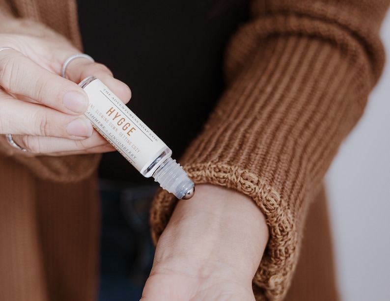 Puede incluir: Un frasco de vidrio transparente con una bola de rodillo de metal. El frasco est&aacute; etiquetado con "HYGGE" y "ALL-NATURAL DRY SETTING OIL" con los ingredientes "AROMATHERAPY - LINGONBERRY".