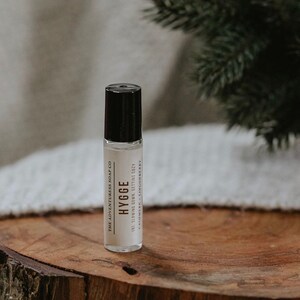 Puede incluir: Un frasco de vidrio transparente con un tap&oacute;n negro. El frasco est&aacute; etiquetado con "HYGGE" y "THE ADVENTURESS SOAP CO". La etiqueta tambi&eacute;n incluye el texto "(NI. SLOWING DOWN. GETTING COZY CASHMERE - LINGONBERRY". El frasco est&aacute; sobre una superficie de madera.