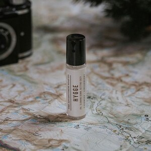 Puede incluir: Un frasco de roll-on blanco con una tapa negra y una etiqueta que dice "THE ADVENTURESS SOAP CO HYGGE (N) SLOWING DOWN. GETTING COZY CASHMERE - LINGONBERRY" en un mapa.