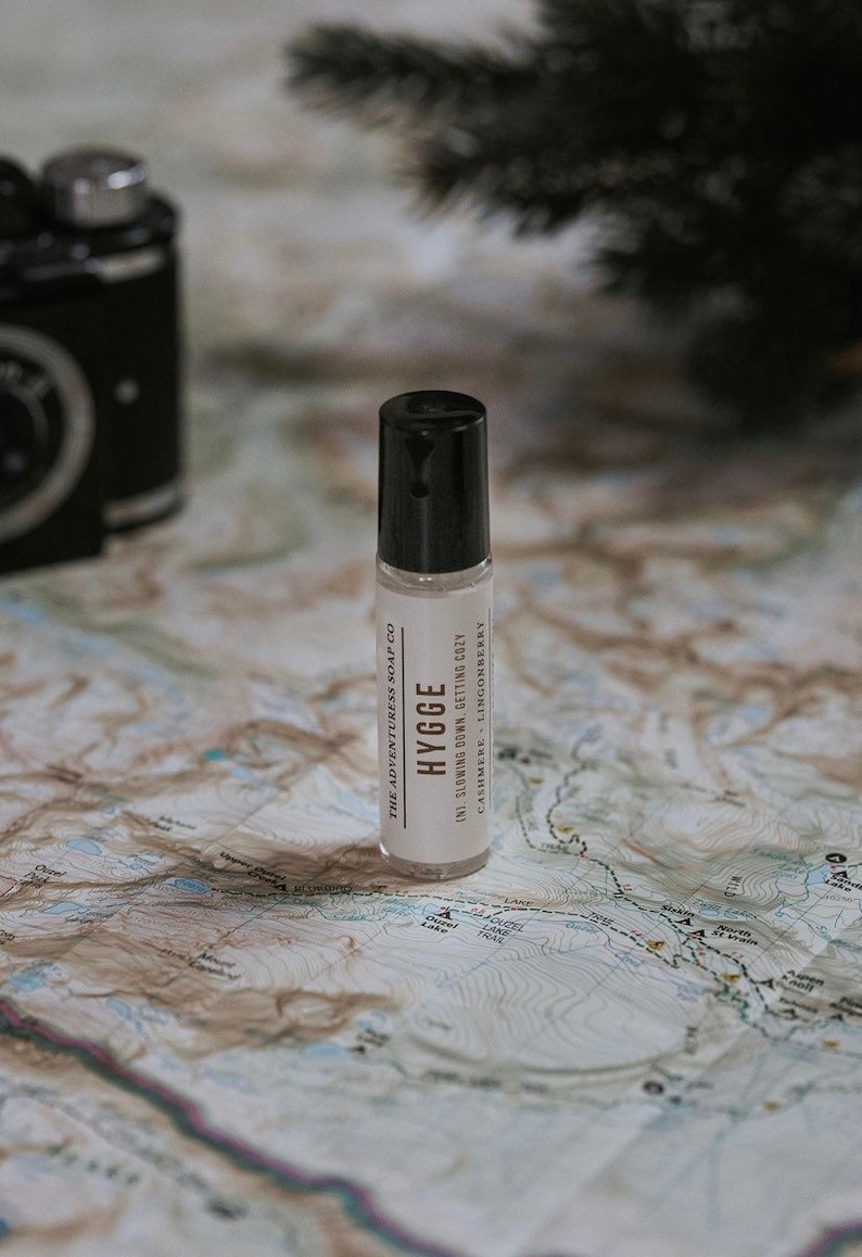 Puede incluir: Un frasco de roll-on blanco con una tapa negra y una etiqueta que dice "THE ADVENTURESS SOAP CO HYGGE (N) SLOWING DOWN. GETTING COZY CASHMERE - LINGONBERRY" en un mapa.