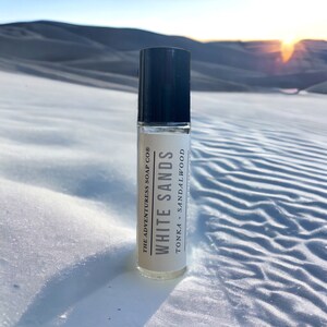 Puede incluir: Un frasco de vidrio transparente con un tap&oacute;n negro. El frasco est&aacute; etiquetado con "White Sands" con el texto "The Adventureress Soap Co." y "Tonka + Sandalwood".