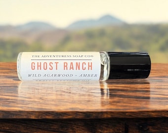 Pode incluir: Um frasco de óleo de perfume de vidro transparente com uma tampa preta repousa sobre uma superfície de madeira. O rótulo diz "Ghost Ranch" com "Wild Agarwood + Amber" abaixo. O fundo apresenta uma paisagem desfocada.