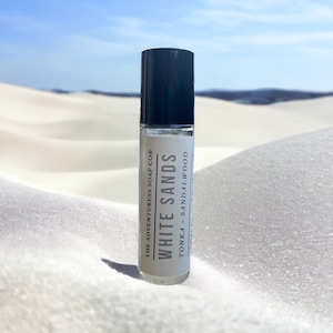 Puede incluir: Un frasco de vidrio transparente con un tap&oacute;n negro. El frasco est&aacute; etiquetado con "White Sands" con el texto "The Adventureress Soap Co." y "Tonka + Sandalwood". El frasco est&aacute; sobre una superficie de arena blanca.