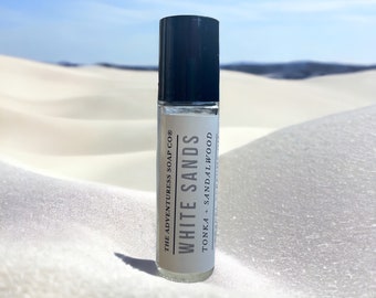 Pode incluir: Um frasco de vidro transparente com uma tampa preta. O frasco é etiquetado com "White Sands" com o texto "The Adventureress Soap Co." e "Tonka + Sandalwood". O frasco está sobre uma superfície de areia branca.