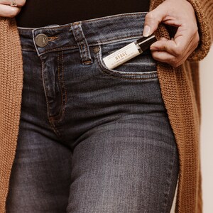 Puede incluir: Una persona lleva pantalones vaqueros azules con una peque&ntilde;a botella de aceite esencial en el bolsillo delantero. La botella tiene una etiqueta blanca con la palabra "HYGGE" impresa en ella.