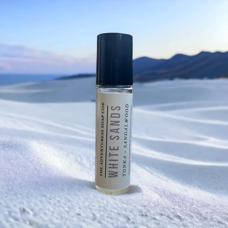 Puede incluir: Un frasco de vidrio transparente con un tap&oacute;n negro y una etiqueta blanca. La etiqueta dice "White Sands" y "The Adventureress Soap Co." con el texto "Tonka + Sandalwood" debajo. El frasco est&aacute; sobre una superficie blanca.