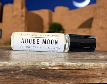 Pode incluir: Um frasco de perfume roll-on de vidro transparente com tampa preta, rotulado "Adobe Moon" com texto "Blackberry + Saffron". O frasco repousa sobre uma superfície de madeira, com um céu noturno e uma lua crescente ao fundo.