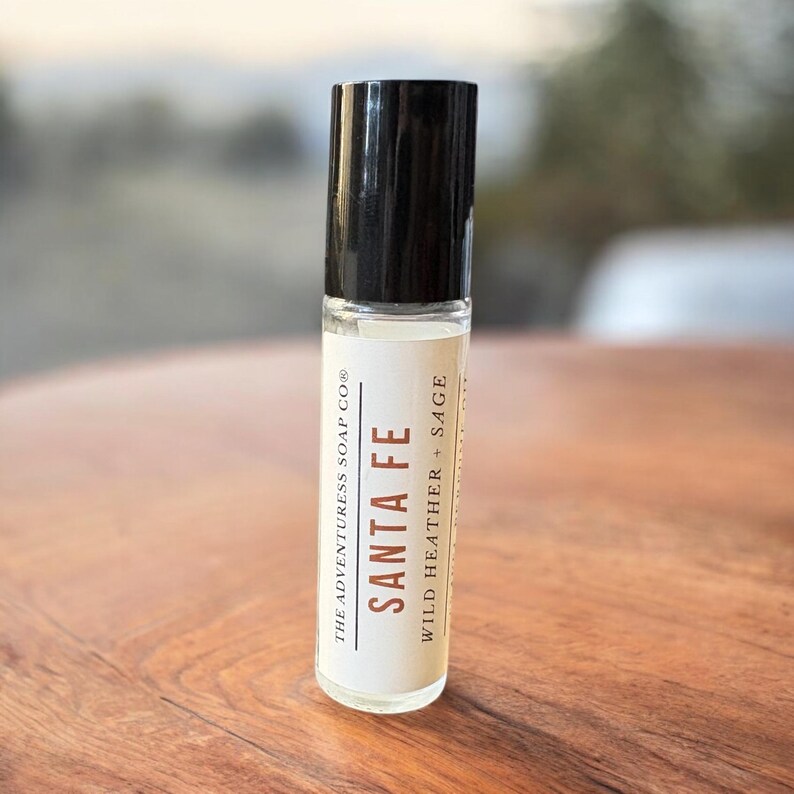 Pode incluir: Um frasco roll-on de vidro transparente com tampa preta. O r&oacute;tulo diz "SANTA FE" com "WILD HEATHER + SAGE" abaixo. O frasco &eacute; de "THE ADVENTURESS SOAP CO&reg;". O frasco est&aacute; sobre uma superf&iacute;cie de madeira.