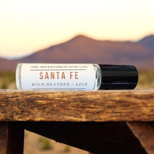 Pode incluir: Um frasco de &oacute;leo de perfume de viagem de vidro transparente com uma tampa preta, rotulado "SANTA FE" com o texto "WILD HEATHER + SAGE". O frasco repousa sobre uma superf&iacute;cie de madeira com um fundo desfocado de montanha des&eacute;rtica.