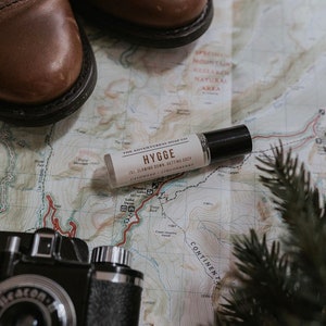 Puede incluir: Una peque&ntilde;a botella de vidrio de aceite esencial con una tapa negra. La botella est&aacute; etiquetada "The Adventureress Soap Co. Hygge Ini. Slowing Down, Getting Cozy Cashmere Lingonberry". La botella est&aacute; sobre un mapa con una c&aacute;mara y botas de cuero marr&oacute;n.