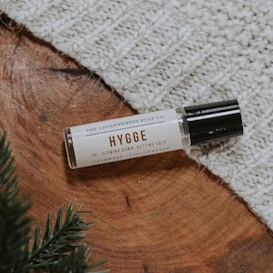 Puede incluir: Una peque&ntilde;a botella blanca con una tapa negra y una etiqueta que dice "THE ADVENTURESS SOAP CO HYGGE (N). SLOWING DOWN. GETTING COZY CASHMERE LINGONBERRY". La botella est&aacute; sobre una superficie de madera con una manta de punto blanca en el fondo.