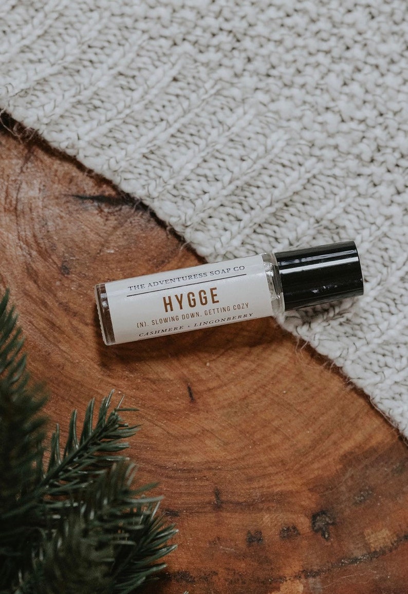 Puede incluir: Una peque&ntilde;a botella blanca con una tapa negra y una etiqueta que dice "THE ADVENTURESS SOAP CO HYGGE (N). SLOWING DOWN. GETTING COZY CASHMERE LINGONBERRY". La botella est&aacute; sobre una superficie de madera con una manta de punto blanca en el fondo.