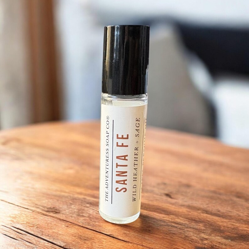 Pode incluir: Um frasco roll-on de vidro transparente com tampa preta. O r&oacute;tulo diz "SANTA FE" com "WILD HEATHER + SAGE" abaixo. O frasco est&aacute; sobre uma superf&iacute;cie de madeira. O texto "THE ADVENTURESS SOAP CO&reg;" tamb&eacute;m est&aacute; no r&oacute;tulo.