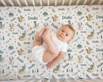 May include: A baby lies on a crib sheet with a repeating pattern of dragons, stars, and the name "Drake" in a whimsical font. The sheet features a soft color palette of white, gold, and teal, creating a fantasy theme.