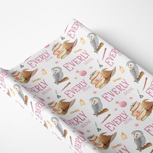 May include: White changing pad cover with a repeating pattern of watercolor illustrations. The design includes a brown wizard hat, a gray owl, a wand, a potion bottle, and the name "EVERLY IRENE" in pink.