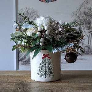 Könnte beinhalten: Ein weißer Keramikbecher mit gesprenkelter Oberfläche und einem grünen Weihnachtsbaum-Motiv, das von einer roten Schleife gekrönt wird. Der Becher ist mit weißen Rosen, grünem Blattwerk, weißen Beeren, silbernen Glitzerblättern und einer Metallglocke gefüllt.