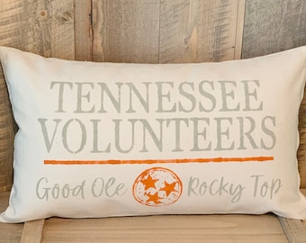 May include: A white pillow with the words "Tennessee Volunteers" in gray text, a thin orange line, and the words "Good Ole Rocky Top" in gray text. A stylized orange star with three white stars inside is centered on the pillow.