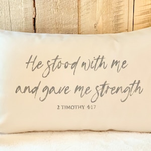 May include: A rectangular white pillow with the text "He stood with me and gave me strength" in gray script. The Bible verse "2 Timothy 4:17" is below the text. The pillow is on a light-colored surface.