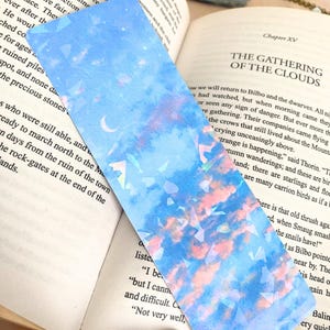 May include: A rectangular bookmark with a dreamy sky design. The bookmark features a blue background with pink and white clouds, a crescent moon, and iridescent geometric shapes. The bookmark is placed on an open book.
