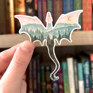 May include: A dragon-shaped sticker with a watercolor design. The wings are pink and the body features a forest scene in shades of green and blue. A crescent moon is visible on one wing. The sticker is held in a hand.