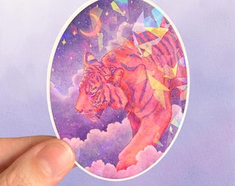May include: Oval sticker featuring a watercolor illustration of a pink and orange tiger walking through a dreamy, purple and pink cloudy sky with a crescent moon and stars. The tiger has dark stripes and is surrounded by colorful geometric shapes.