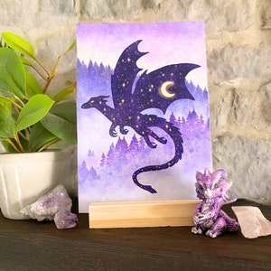 May include: A fantasy art print of a dragon silhouetted against a purple forest and sky filled with stars and a crescent moon. A small purple dragon figurine sits on a shelf next to the print. The print is displayed on a wooden stand.