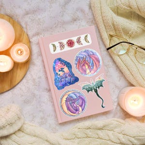 May include: A pink journal adorned with fantasy-themed stickers. The stickers feature dragons, moon phases, and a forest scene. The journal is surrounded by candles, a cozy blanket, and eyeglasses.
