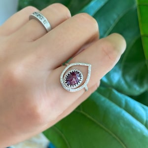May include: A silver ring with a teardrop-shaped design, featuring a central oval-cut, deep red gemstone. The ring is encrusted with small, clear stones along the teardrop outline. Another silver band ring is also visible.