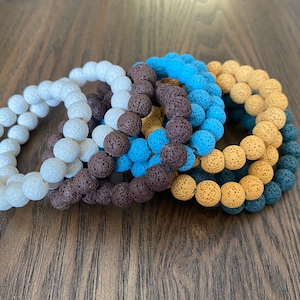 Könnte beinhalten: Eine Sammlung von Armbändern mit Perlen in verschiedenen Farben: Weiß, Braun, Blau und Beige. Jedes Armband besteht aus kleinen, runden Perlen mit strukturierter Oberfläche. Die Armbänder sind gestapelt und zeigen die verschiedenen Farboptionen.