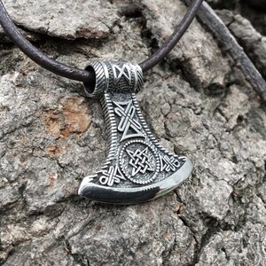Puede incluir: Collar con colgante de hacha vikinga de plata con intrincados diseños de nudos y un cordón de cuero.