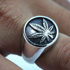 Könnte beinhalten: Ein silberner Ring mit einer runden, schwarzen Oberfläche, die mit einem erhabenen, silbernen Marihuana-Blatt verziert ist.