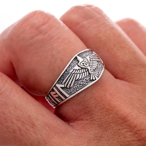 Puede incluir: Un anillo plateado con un diseño detallado de cuervo y un nudo celta. El anillo tiene texto grabado en el lateral. El cuervo tiene las alas extendidas. El anillo se lleva en un dedo.