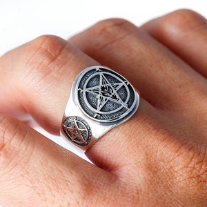 Peut inclure: Une bague en argent avec un motif de pentagramme représentant une tête de chèvre au centre. La bague est portée au doigt d'une personne.