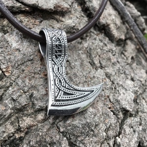 May include: A silver axe pendant with intricate knotwork and a detailed design. The pendant is hanging from a brown leather cord.