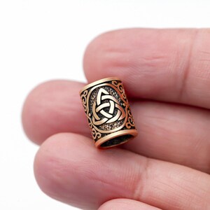 May include: A gold-coloured metal bead with a Celtic knot design. The bead is textured and has a detailed pattern.