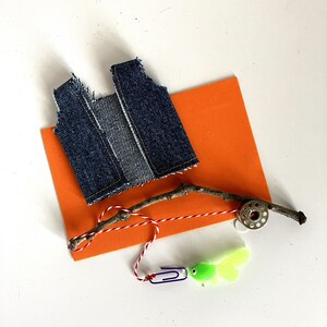 May include: A blue denim vest, a twig with red and white string, a spool of thread, and a green and yellow fish lure on an orange background.