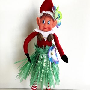 May include: A Christmas elf doll wearing a red and white outfit, a green grass skirt, and a flower crown. The elf has blue eyes and a smiling face.