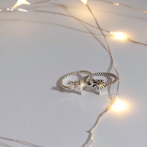 May include: Two silver rings with palm tree designs. The rings are on a white surface with string lights in the background.