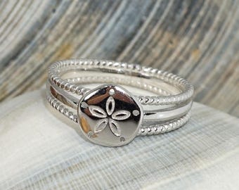 Op de afbeelding: Een zilveren ring met een centrale ronde bedel voorzien van een bloemmotief met vijf bloemblaadjes. De ringband bestaat uit twee delen: een gladde binnenring en een buitenring met een korrelige, getextureerde afwerking rondom de bedel.