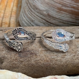 May include: Two silver rings made from vintage silverware, displayed on a piece of driftwood with seashells.