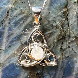 May include: A silver-toned pendant necklace featuring a Celtic knot design with crescent moon accents. The pendant has a central, round, light-colored stone. The necklace chain is delicate and silver. The background is a mottled, blue-gray surface.